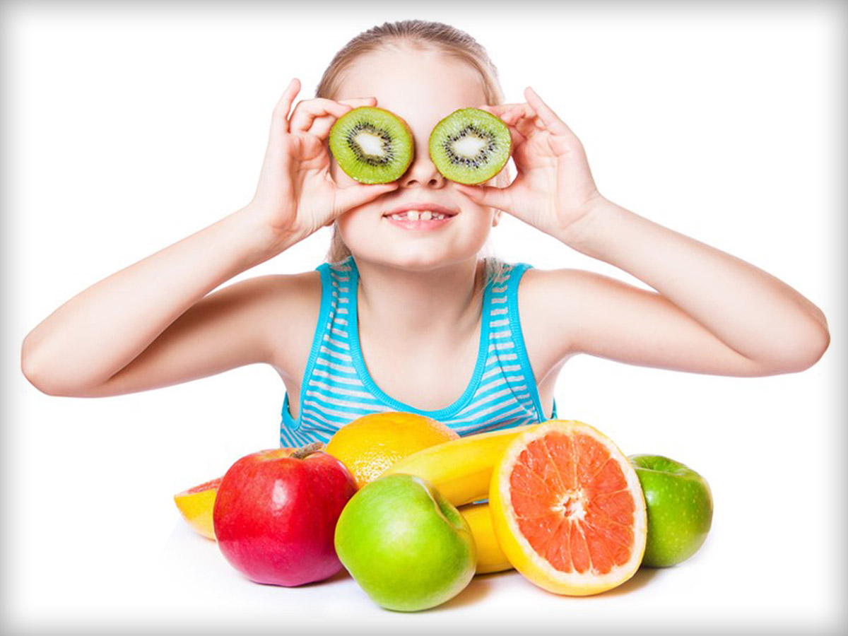 Girl with a variety of fruits in front of her, holding two kiwi slices in front of her eyes